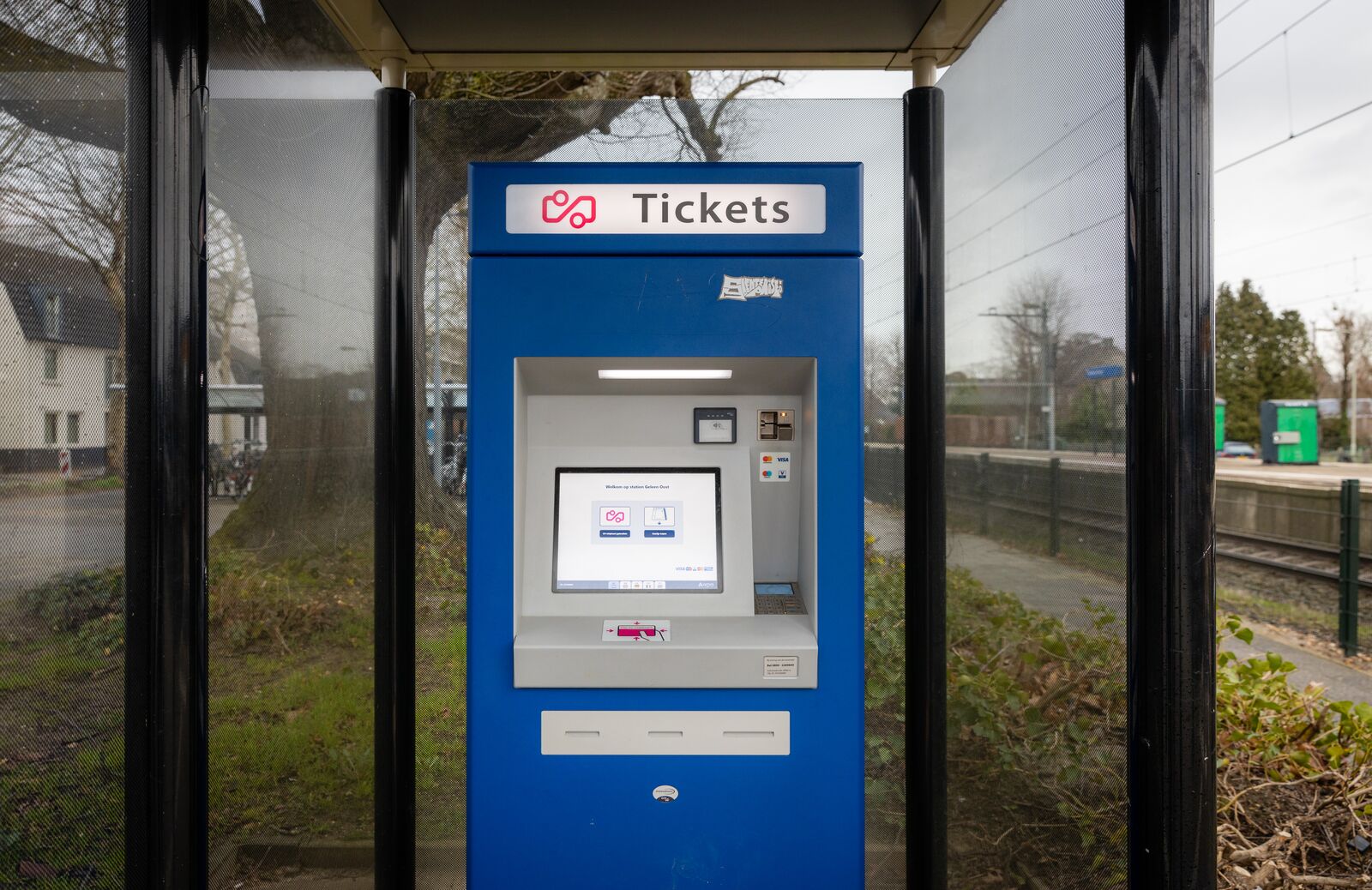 Blauwe ticketautomaat met touchscreen onder een overkapping op een treinperron.
