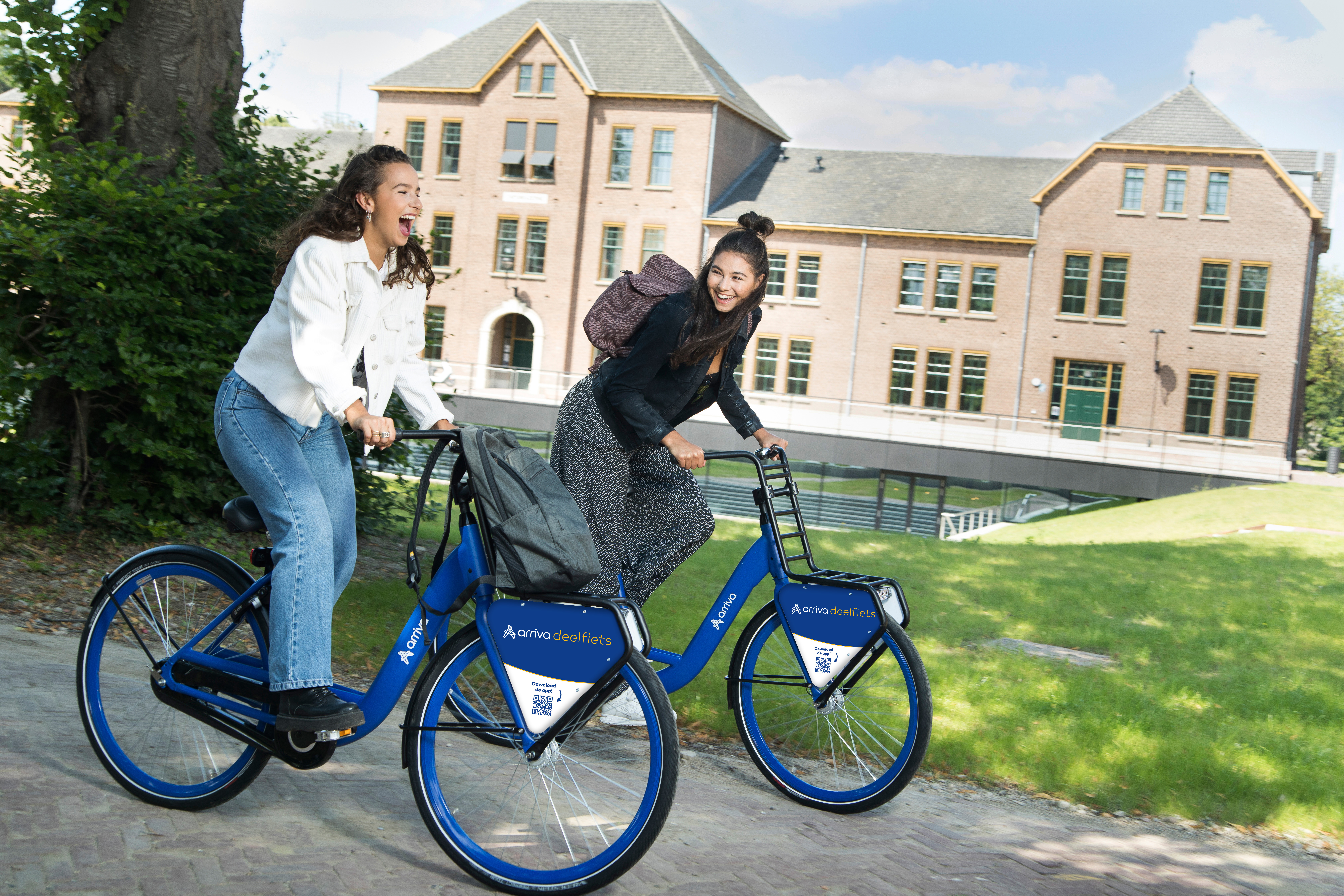 Twee lachende studentes fietsen op blauwe Arriva-deelfietsen door een groene omgeving.