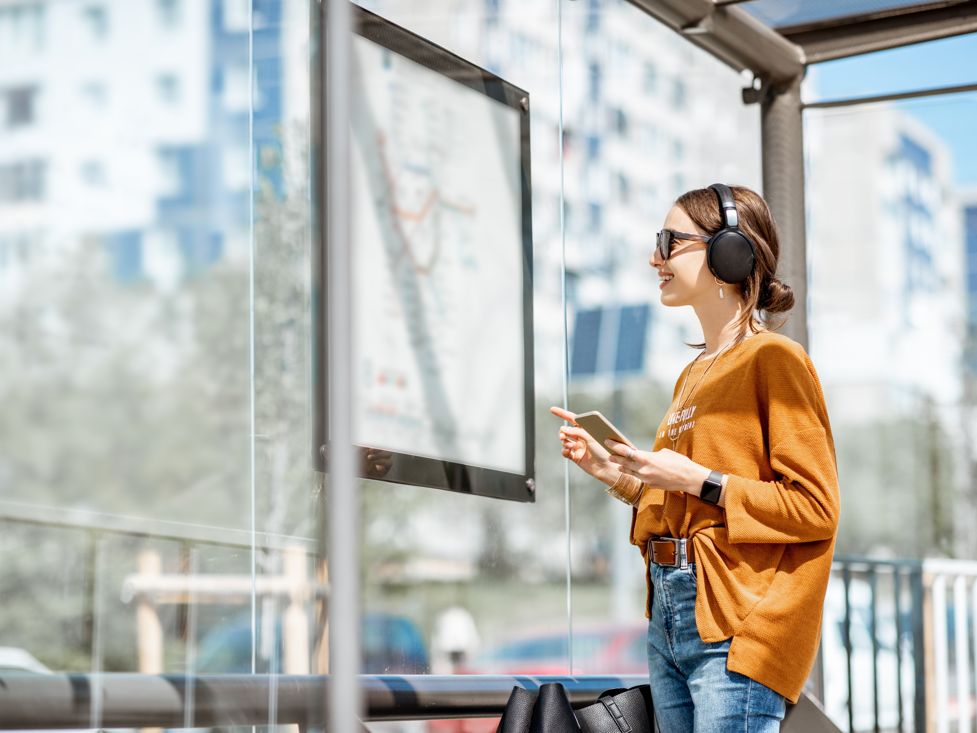 Jonge vrouw staat op stralende dag met koptelefoon lachend te kijken naar een vertrekstaat op een bushalte.