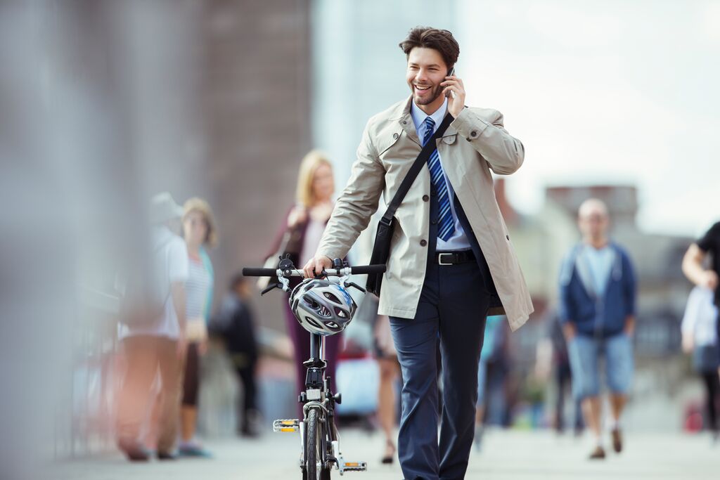 Een bellende, lachende man in business-casual outfit loopt met vouwfiets en helm aan de hand door een stad.