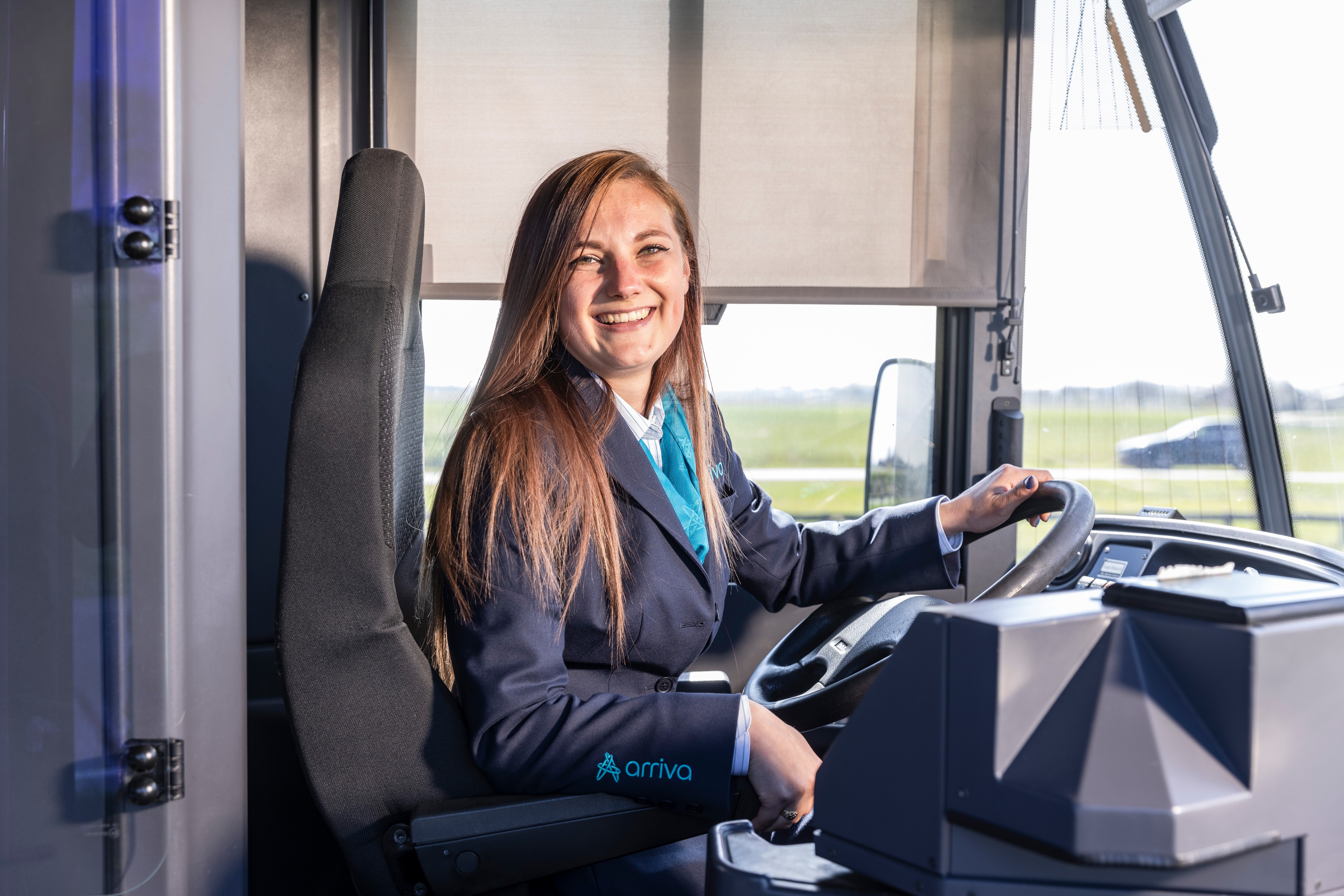 Vrouwelijke buschauffeur met lang haar en in uniform zit lachend achter het stuur van haar Arriva-bus.