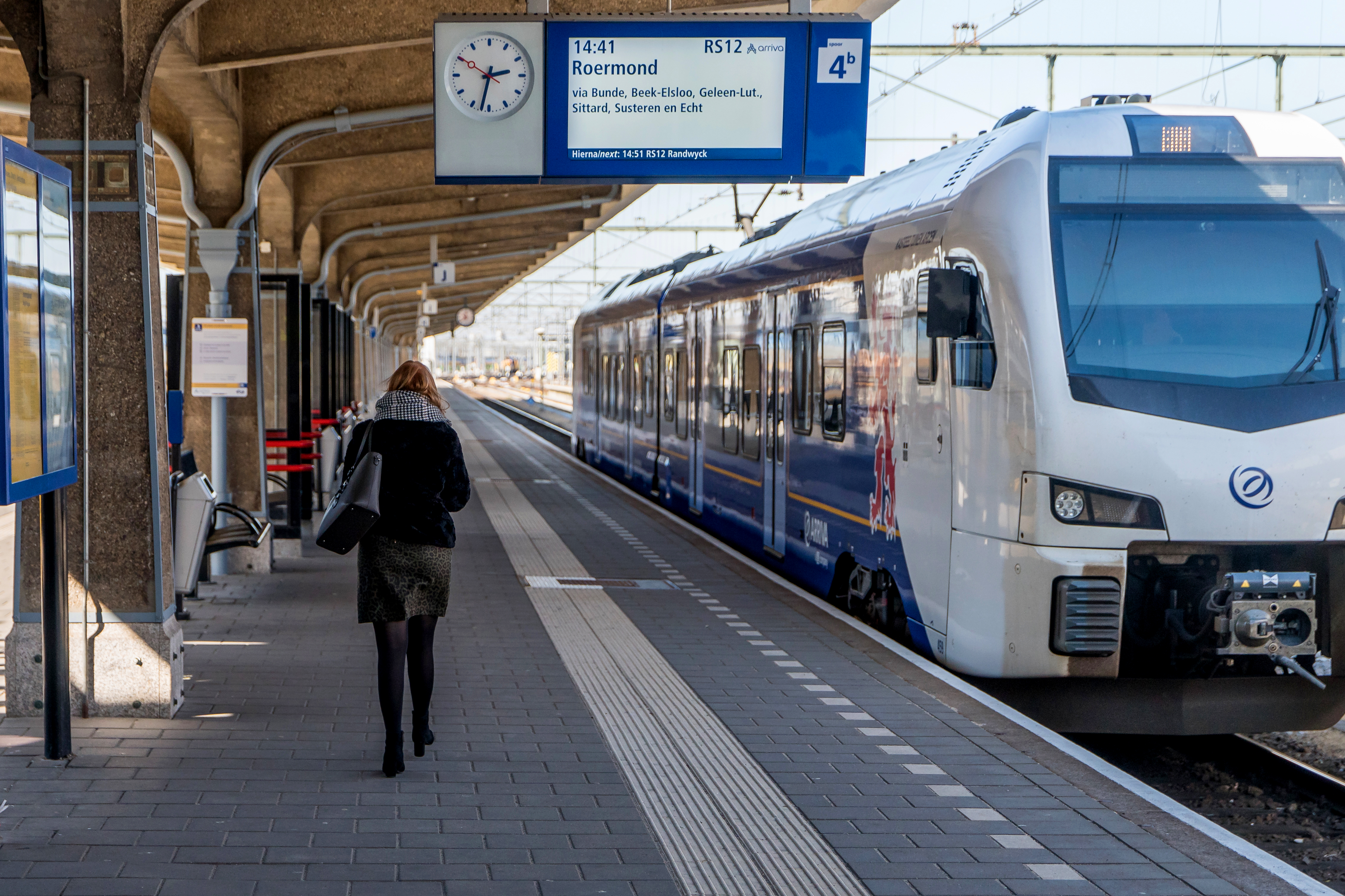 Reiziger loopt over perron van een station langs een blauwwitte trein van Arriva die naar Roermond gaat.