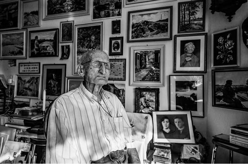 Een zwart-witfoto met daarop een heel oude man in overhemd die in de verte staart, de muur achter hem is volgehangen met foto's.